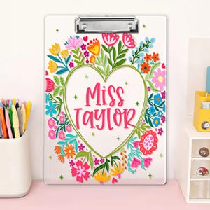 May include: A white clipboard with a silver clip, decorated with a floral heart design. The heart contains the text "Miss Taylor" in pink. The floral arrangement includes pink, yellow, orange, and blue flowers with green leaves.