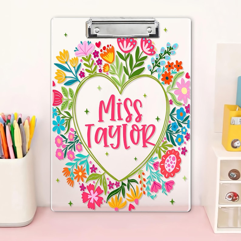 May include: A white clipboard with a silver clip, decorated with a floral heart design. The heart contains the text "Miss Taylor" in pink. The floral arrangement includes pink, yellow, orange, and blue flowers with green leaves.