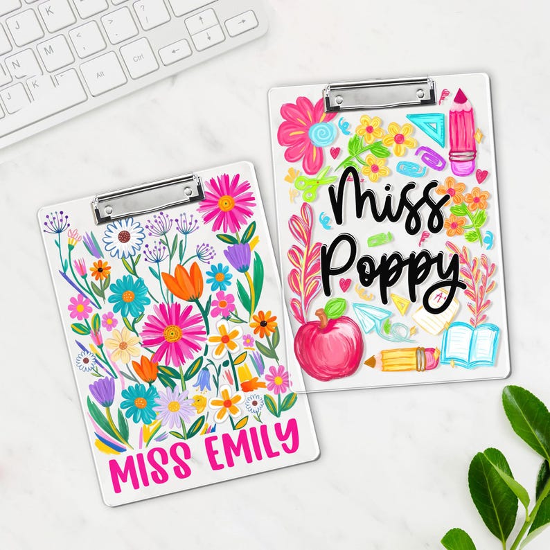 May include: Two white clipboards with floral designs. One is labelled "Miss Emily" in pink, featuring colourful flowers. The other, "Miss Poppy," has an apple, pencil, and book illustrations. Both have silver clips.