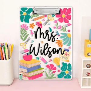 May include: A clear clipboard with a metal clip, decorated with colourful floral and school-themed illustrations. The name "Mrs. Wilson" is written in black script. Includes images of pencils, scissors, books, and an apple.