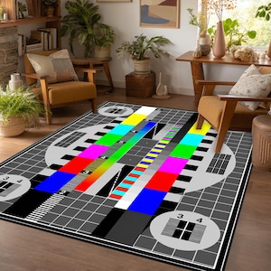 Op de afbeelding: Een rechthoekig vloerkleed met een televisie-testbeeldontwerp. Het kleed toont een raster van grijze vierkanten, cirkelvormige elementen met zwart-witte lijnpatronen en een centrale diagonale band van levendige primaire en secundaire kleuren.