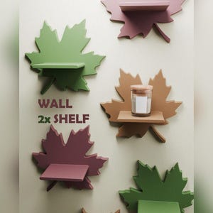 May include: A collection of wall-mounted shelves shaped like maple leaves in shades of green, brown, and maroon. The text WALL 2x SHELF is printed on the surface. One shelf holds a small glass candle with a wooden lid and a blank white label.