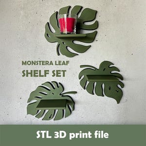Può includere: Un set di tre mensole da parete color verde oliva a forma di foglie di monstera. Le mensole presentano dettagli intagliati e una superficie orizzontale piatta. Una mensola sostiene un portacandele in vetro trasparente con una candela rosa acceso. Testo: MONSTERA LEAF SHELF SET e STL 3D print file.