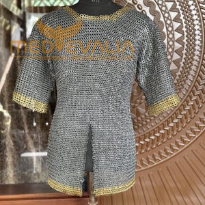 Peut inclure: Un maillot de cotte de mailles argenté avec des manches courtes et une bordure dorée autour de l'encolure et des poignets. Le maillot est présenté sur un mannequin noir. Le texte "MEDIEVALIA" est visible sur le côté gauche.