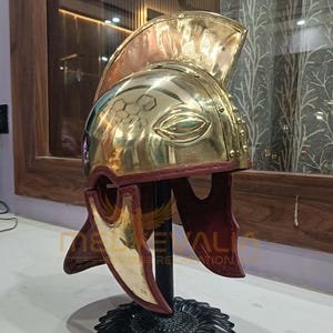 May include: A polished brass helmet with a prominent crest and cheek guards. The helmet features an eye-shaped design and a burgundy interior. It is displayed on a black stand, showcasing its historical design and craftsmanship.