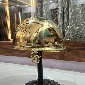 Könnte beinhalten: Ein polierter Messinghelm mit abgerundeter Oberseite und dekorativem Rand. Der Helm ist auf einem schwarzen Ständer mit einem verzierten Sockel montiert. Die Oberfläche des Helms reflektiert die Umgebung.