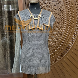Puede incluir: Una camisa de cota de malla sobre un maniquí. La camisa está hecha de pequeños anillos de metal entrelazados. Tiene un cuello y detalles en los hombros con adornos dorados. El maniquí está sobre una superficie de madera.