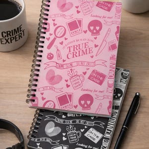 May include: Three spiral-bound notebooks with true crime designs. The top one is pink with crime scene graphics and the text "TRUE CRIME." The second notebook is black and white with similar graphics. A pen and headphones are also visible.
