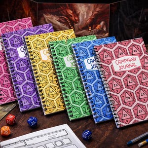May include: A selection of spiral-bound notebooks with covers in various colours, each displaying a repeating pattern of a twenty-sided die. The notebooks are pink, purple, yellow, green, and blue. One notebook has the words "Campaign Journal".
