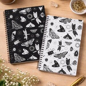 May include: Two insect-themed notebooks with spiral bindings. One has a black cover with white insects, the other a white cover with black insects. A gold pen and a coffee cup are also visible on a wooden surface.