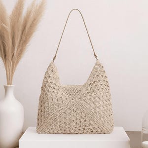 May include: A beige crochet shoulder bag with a long strap. The bag has a geometric open weave design and a neutral-colored lining. The bag is displayed on a white surface.
