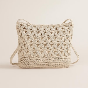 May include: A beige, woven crossbody bag with a floral pattern on the front and a solid, woven bottom. The bag has two long, braided straps and a neutral-colored interior. The bag is made of a natural fiber.