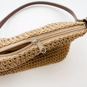Pu&ograve; includere: Primo piano di una borsa di paglia intrecciata con un cinturino in pelle marrone. La borsa presenta una cerniera beige e una trama intrecciata. La borsa &egrave; di colore beige chiaro.