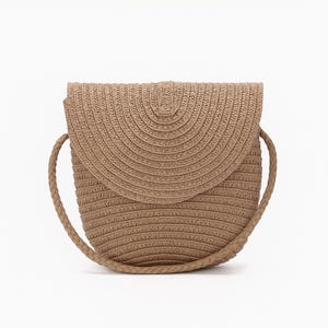 May include: A tan-coloured, woven straw crossbody bag with a rounded flap and braided shoulder strap. The bag has a circular shape and a woven texture, perfect for carrying essentials. The bag is set against a white background.