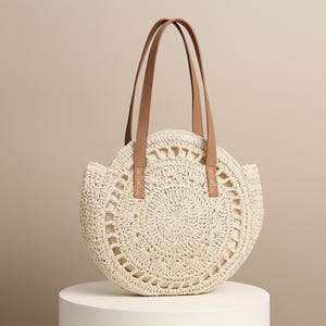 May include: Cream-coloured, round woven tote bag with brown leather handles. The bag features a detailed open weave design and a light-coloured interior. The handles are attached with small, square leather patches.