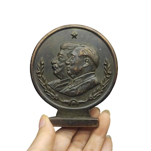 Puede incluir: Una placa metálica circular de tono bronce con retratos en relieve de dos hombres de perfil, colocados uno detrás del otro. Una pequeña estrella de cinco puntas está en la parte superior y ramas de laurel enmarcan los lados inferiores.