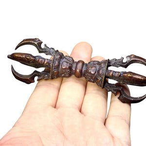 May include: A dark bronze-coloured metal vajra ritual object featuring a central grip with intricate relief carvings and two symmetrical ends, each composed of four curved prongs extending from a central point, creating a balanced design.