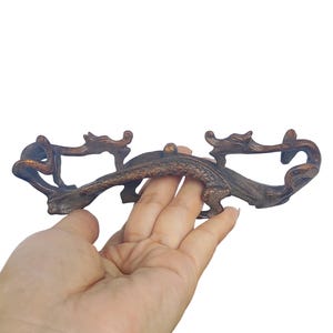 Op de afbeelding: Een donkerbruine, gesneden houten displaystandaard met ingewikkelde, langwerpige draakachtige figuren aan beide uiteinden. De standaard heeft een gebogen, getextureerde centrale brug en verhoogde steunen, met een gepolijste, diepe houtnerf.