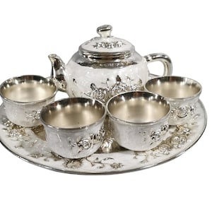 May include: A silver and white tea set comprising a teapot, four small cups, and a round serving tray. Each piece features a raised floral rose pattern with a metallic silver finish on the rims and handles, contrasted by white enamel detailing.
