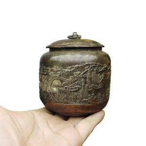 Peut inclure: Un petit pot en métal de couleur bronze foncé, de forme ronde, doté d'un couvercle amovible avec un petit anneau sur le dessus. La surface extérieure est ornée d'un paysage en relief représentant des bateaux, de l'eau et des bâtiments.