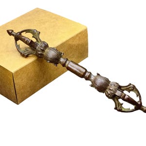 Peut inclure: Un objet rituel vajra en métal de couleur bronze doté d'un manche cylindrique central flanqué de deux sections symétriques en forme de lotus et d'extrémités à griffes. L'objet présente une finition patinée sombre et vieillie.