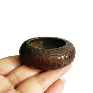 May include: A bronze-colored, circular ring with an embossed pattern of swirling designs. The ring has a hollow center and appears to be made of metal. The ring is held in a hand against a white background.