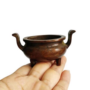 Puede incluir: Un pequeño quemador de incienso antiguo de bronce con tres patas y dos asas curvas. El recipiente tiene un cuerpo redondeado y una amplia abertura. El bronce tiene una pátina oscura y envejecida, lo que sugiere su antigüedad. El quemador se sostiene en una mano sobre un fondo blanco.