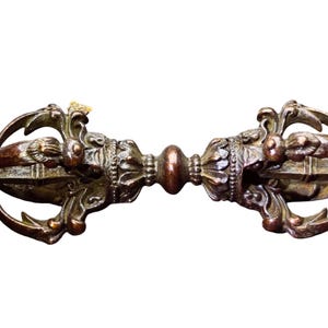 Peut inclure: Un objet rituel vajra en métal de couleur bronze, doté d'une poignée centrale et de deux extrémités symétriques aux pointes incurvées. La surface présente une patine sombre et usée avec des sculptures en relief et des motifs floraux.
