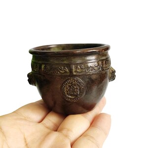 Pode incluir: Um pequeno pote castanho escuro, de estilo antigo, com corpo arredondado e abertura larga. O pote apresenta padrões decorativos em relevo ao redor da borda e um emblema circular na frente. Duas alças de cabeça de leão nas laterais. O pote é segurado em uma mão.