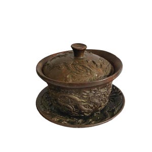 Op de afbeelding: Een bronzen theeservies met ingewikkelde draken- en bloemreliëf. De set bevat een kom met deksel en een bijpassende schotel. Het ontwerp heeft gedetailleerde patronen en een donkere, verouderde afwerking.