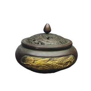 May include: A round, dark bronze incense burner featuring a removable lid with an intricate openwork design. The body of the vessel displays a prominent, raised gold-coloured relief of a mythical bird, complemented by a small, pointed finial on top.