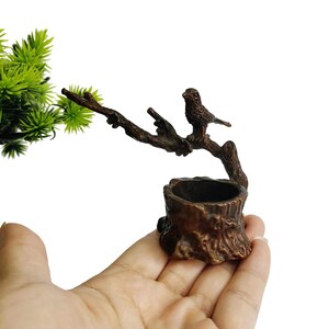 May include: A bronze-colored miniature sculpture of a bird perched on a tree branch, above a tree stump-shaped planter. The sculpture is held in a hand, with green foliage in the background. The bird is detailed with textured feathers.
