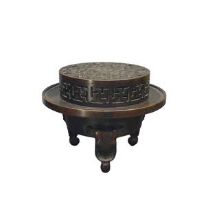 May include: A dark bronze incense burner featuring a circular lid with an intricate openwork pattern and a wide, flat rim. The body of the vessel is supported by three curved legs, and the side of the lid displays a repeating geometric swastika motif.