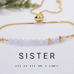 May include: Gold-toned bracelet featuring a string of pale lavender beads and gold accents. The bracelet has an adjustable chain and a sliding clasp. The word "SISTER" is printed below the bracelet, along with a Morse code representation.