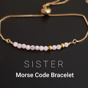 May include: Gold-toned adjustable bracelet with pink and gold beads. The bracelet features the word "SISTER" and the text "Morse Code Bracelet" in white font.