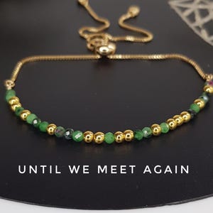 May include: Gold-toned bracelet with a delicate chain and a sliding clasp. The bracelet features a row of small, round, gold beads interspersed with faceted green beads. The text "UNTIL WE MEET AGAIN" is at the bottom.