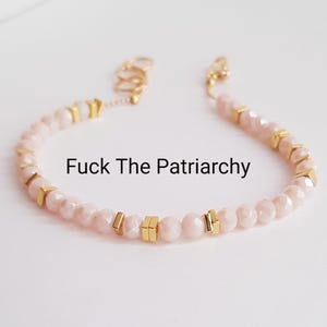 May include: A delicate bracelet featuring pale pink faceted beads and gold-colored geometric accents. The bracelet has the text "Fuck The Patriarchy" in black. The jewelry is on a white background.