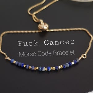 May include: A gold-colored adjustable bracelet with blue and gold beads. The bracelet has the text "Fuck Cancer Morse Code Bracelet" in white. The bracelet is on a black surface.