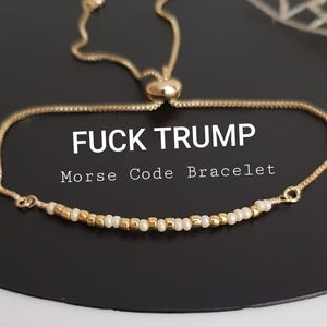 May include: Gold-toned Morse code bracelet with the text "FUCK TRUMP". The bracelet features a delicate chain and a series of small gold and white beads. The adjustable design allows for a comfortable fit. The bracelet is displayed on a black surface.