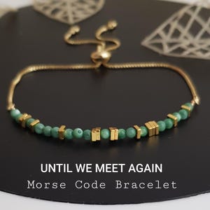 May include: Gold-toned adjustable bracelet with green beads and gold square accents. The bracelet features the text "UNTIL WE MEET AGAIN Morse Code Bracelet" on a black surface. The background has leaf-shaped decorations.