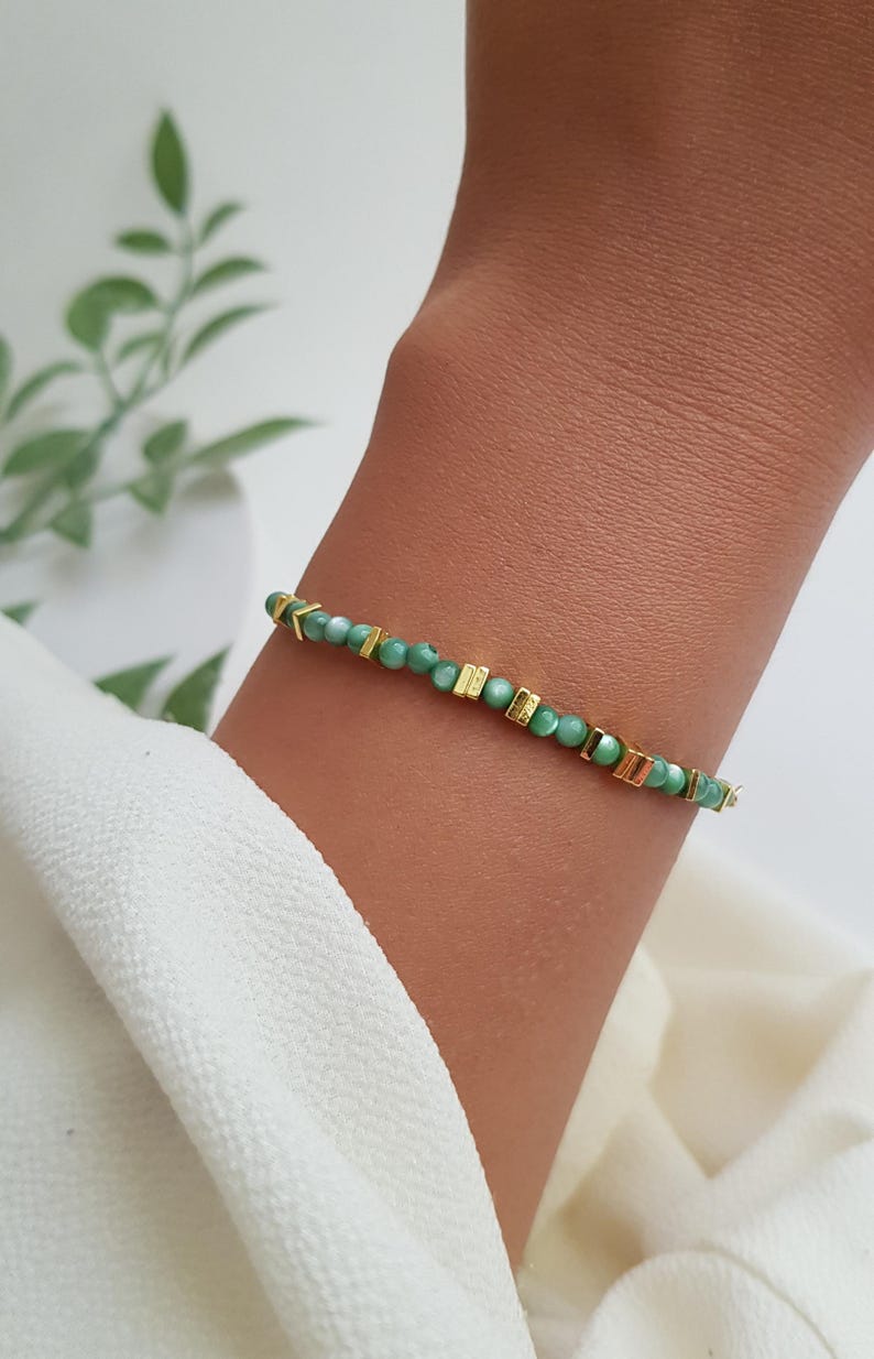 Puede incluir: Una delicada pulsera con peque&ntilde;as cuentas verdes redondas intercaladas con detalles rectangulares y en forma de flecha dorados. La pulsera se lleva en la mu&ntilde;eca, mostrando su dise&ntilde;o y contraste de color. La joya se presenta sobre un fondo neutro.