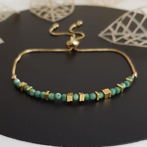 Puede incluir: Una pulsera dorada con cierre deslizante. La pulsera presenta peque&ntilde;as cuentas verdes redondas y cuentas rectangulares doradas. La pulsera se muestra sobre una superficie negra.