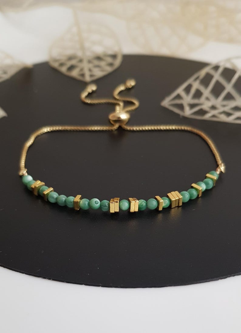 Puede incluir: Una pulsera dorada con cierre deslizante. La pulsera presenta peque&ntilde;as cuentas verdes redondas y cuentas rectangulares doradas. La pulsera se muestra sobre una superficie negra.