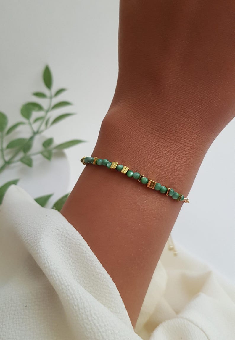 Puede incluir: Una delicada pulsera con peque&ntilde;as cuentas verdes redondas intercaladas con detalles rectangulares dorados. La pulsera est&aacute; ensartada en una fina cadena dorada y se muestra en una mu&ntilde;eca sobre un fondo neutro.