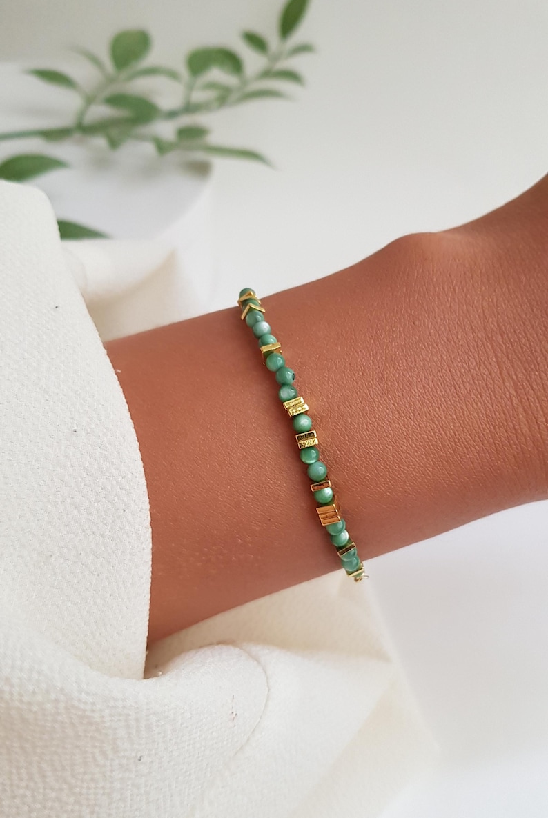 Puede incluir: Una delicada pulsera con peque&ntilde;as cuentas redondas de color verde claro intercaladas con detalles rectangulares y en forma de flecha dorados. La pulsera se muestra en una mu&ntilde;eca, mostrando su dise&ntilde;o y color.