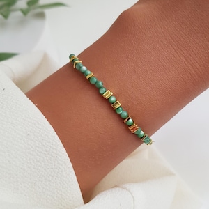 Puede incluir: Una delicada pulsera con peque&ntilde;as cuentas redondas de color verde claro intercaladas con detalles rectangulares y en forma de flecha dorados. La pulsera se lleva en una mu&ntilde;eca, sobre un fondo de tela blanca.