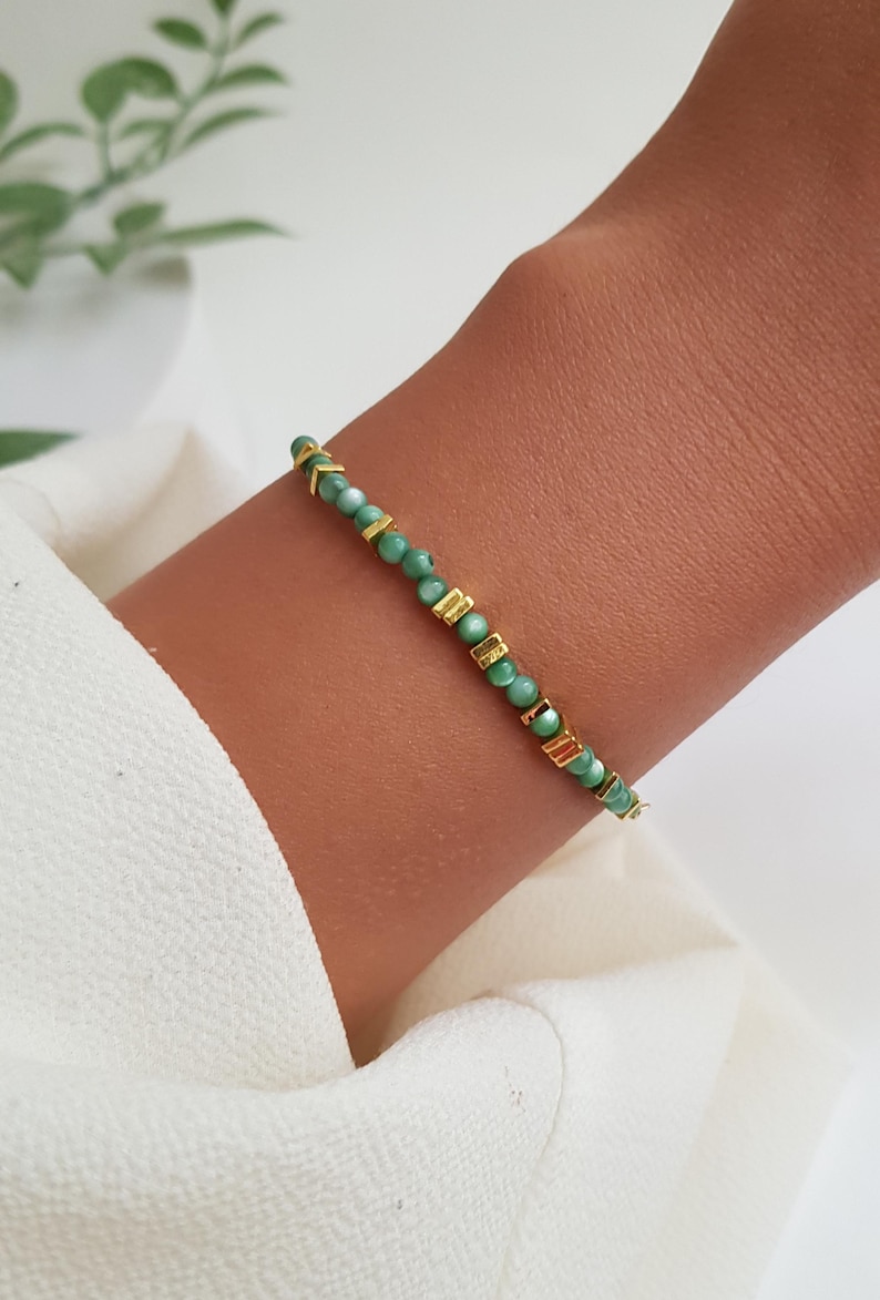 Puede incluir: Una delicada pulsera con peque&ntilde;as cuentas redondas de color verde claro intercaladas con detalles rectangulares y en forma de flecha dorados. La pulsera se lleva en una mu&ntilde;eca, sobre un fondo de tela blanca.