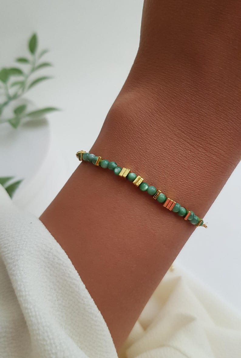 Puede incluir: Una delicada pulsera con peque&ntilde;as cuentas redondas verdes intercaladas con detalles rectangulares dorados. La pulsera est&aacute; ensartada en un fino alambre dorado. La joya se muestra en un brazo.