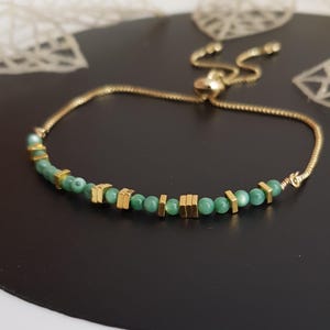 Puede incluir: Pulsera dorada con una delicada cadena y un cierre deslizante para ajustar la talla. La pulsera est&aacute; adornada con peque&ntilde;as cuentas verdes redondas y detalles cuadrados dorados. La joya se exhibe sobre una superficie oscura.