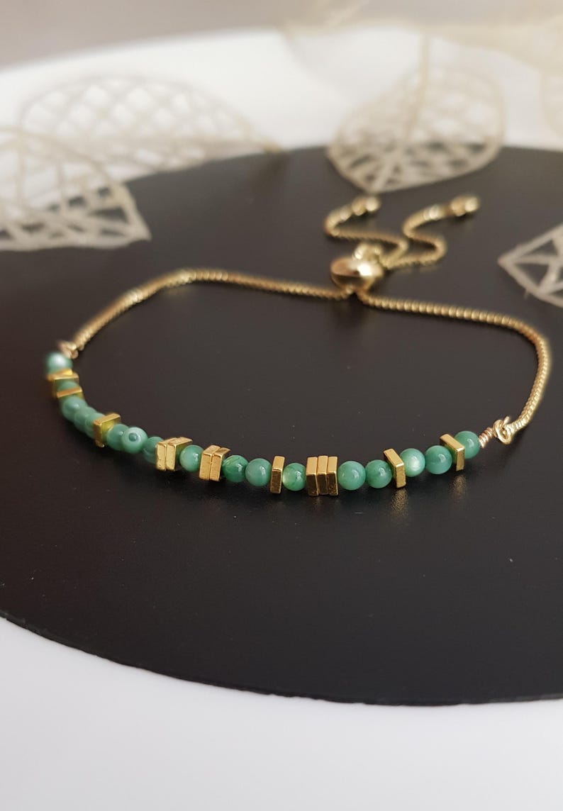 Puede incluir: Pulsera dorada con una delicada cadena y un cierre deslizante para ajustar la talla. La pulsera est&aacute; adornada con peque&ntilde;as cuentas verdes redondas y detalles cuadrados dorados. La joya se exhibe sobre una superficie oscura.
